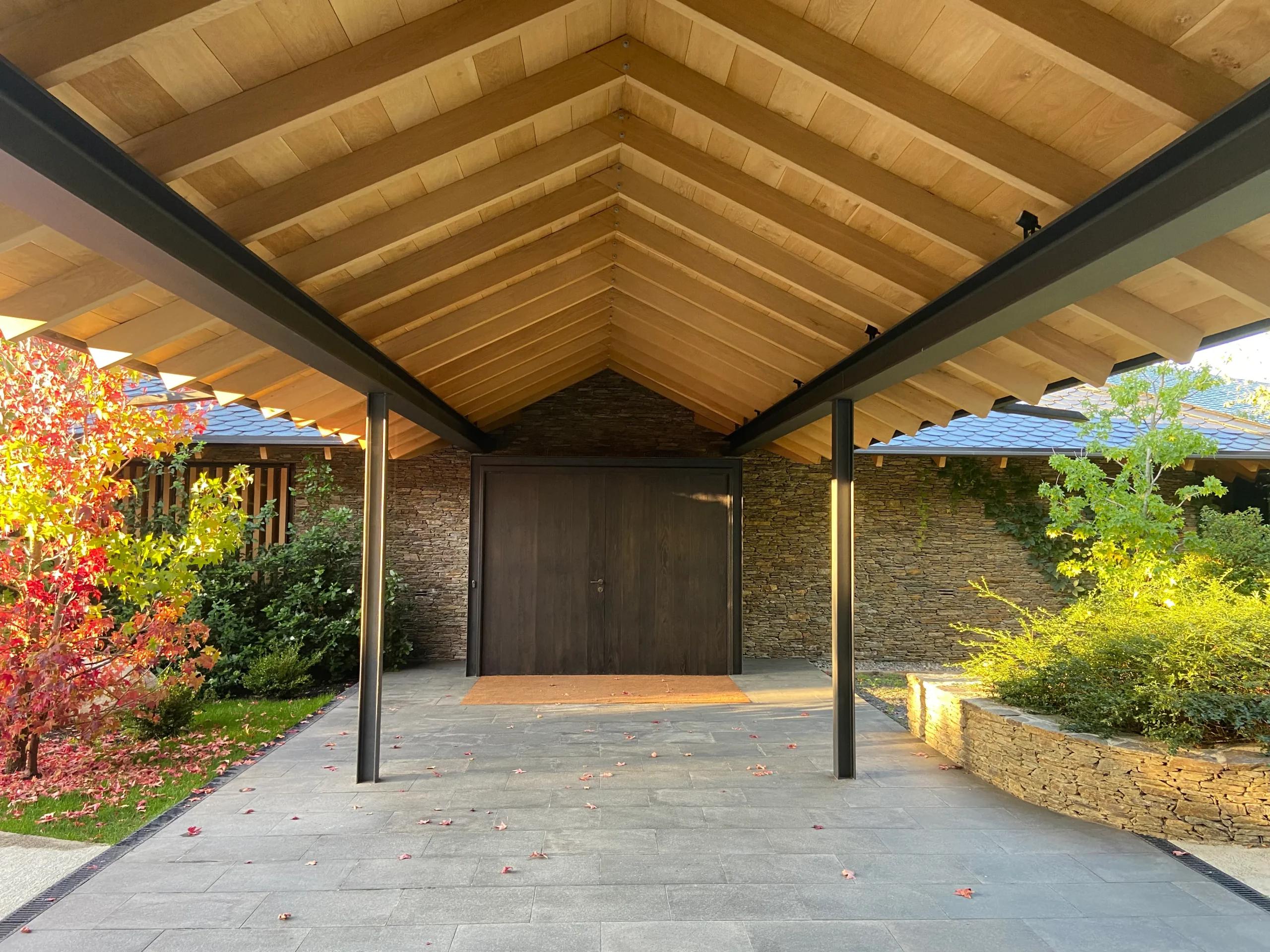 Entrada moderna de casa con porche de madera, paredes de piedra, puerta oscura y vegetación otoñal, ideal para exteriores.