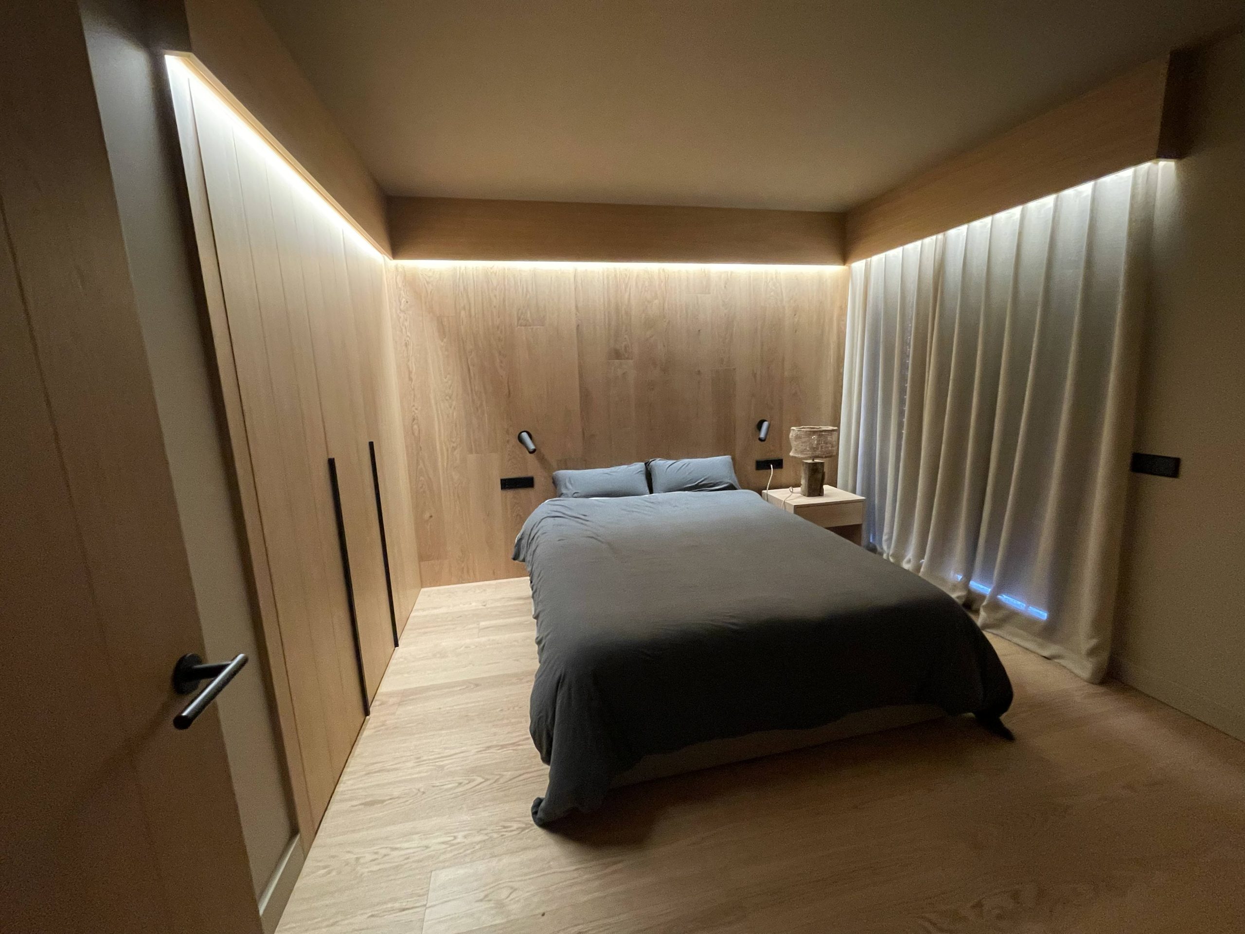 Dormitorio moderno con diseño interior de madera a medida, cama gris, iluminación LED oculta y cortinas claras