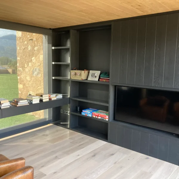 Salón moderno con estanterías negras, televisión integrada, ventanal con vistas a la naturaleza y suelo de madera clara.