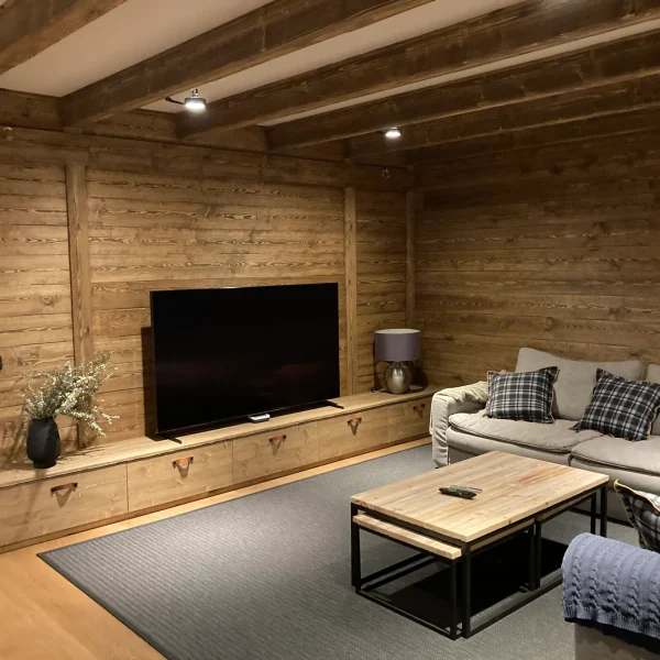 Salón rústico de madera con vigas vistas, sofá gris, mesa de centro de madera y TV, ideal para proyectos de carpintería a medida.