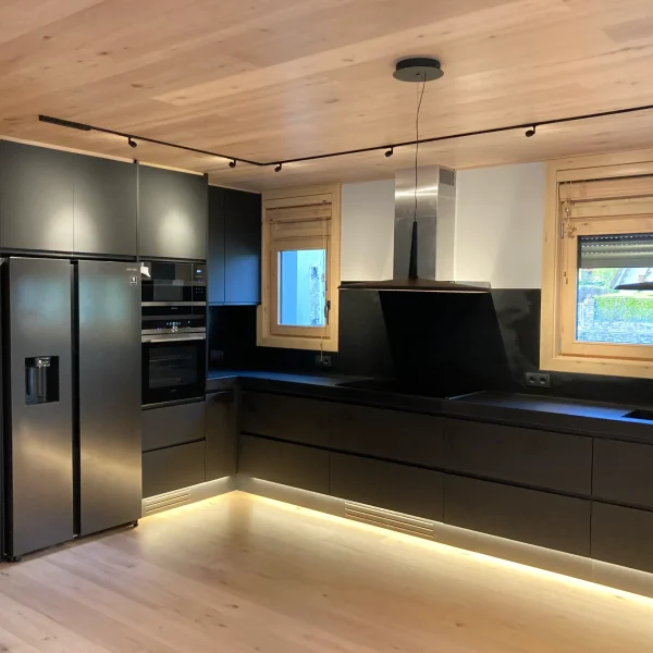 Cocina moderna con gabinetes negros, electrodomésticos de acero inoxidable, iluminación LED y techo de madera clara.