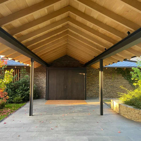 Entrada moderna de casa con porche de madera, paredes de piedra, puerta oscura y vegetación otoñal, ideal para exteriores.