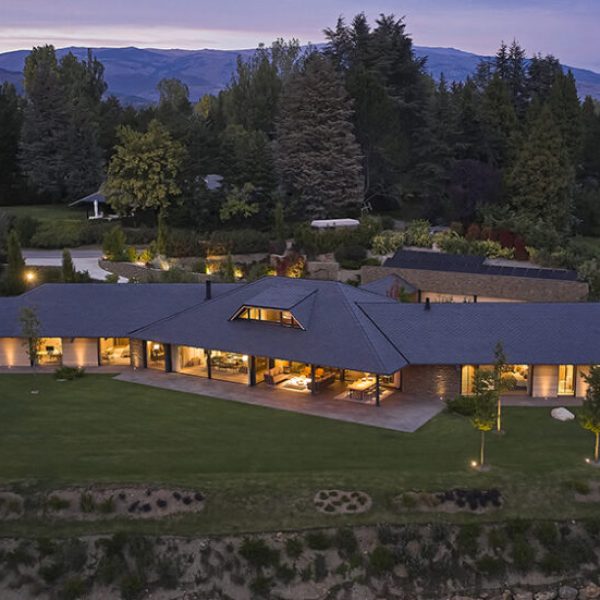 Impresionante casa moderna con techo oscuro y grandes ventanales iluminados al atardecer, rodeada de bosque y montañas.