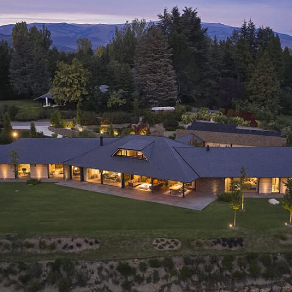 Moderna casa de lujo con tejado oscuro y grandes ventanales iluminados al atardecer, rodeada de bosque y montañas.