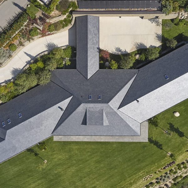 Vista aérea de una casa moderna con tejado de pizarra oscura y diseño arquitectónico en forma de estrella, rodeada de jardine