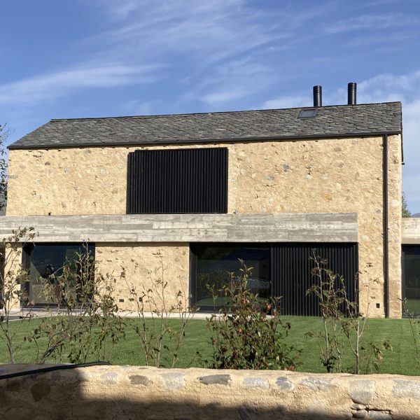 Casa moderna de piedra y hormigón con techo de pizarra, grandes ventanales negros y césped verde bajo cielo azul.