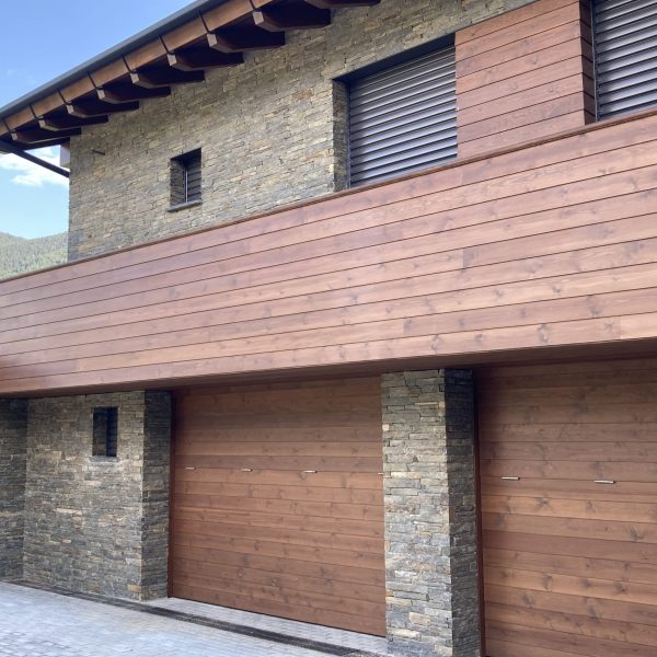 Fachada moderna de casa con revestimiento de madera y piedra, garajes de madera y persianas metálicas, en entorno montañoso.