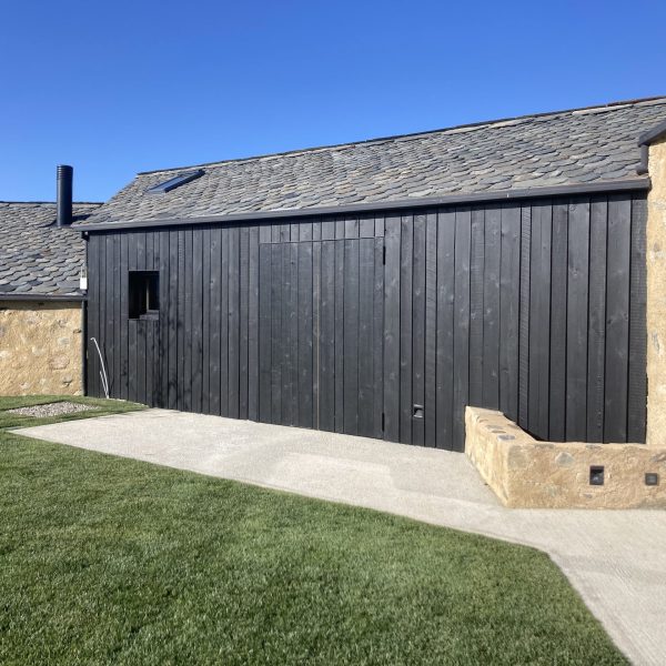 Fachada moderna de casa con revestimiento de madera negra, techo de pizarra y muro de piedra, césped verde y cielo azul.