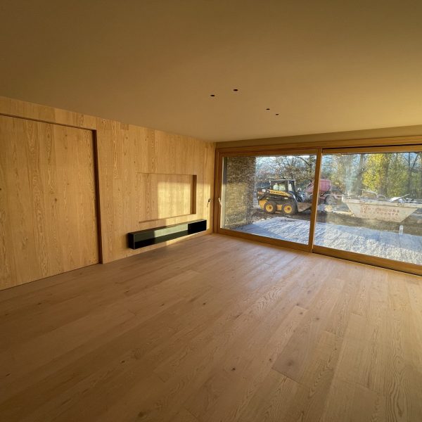 Habitación moderna con paredes y suelo de madera clara, ventana panorámica y estante flotante negro, con vistas a obra exteri