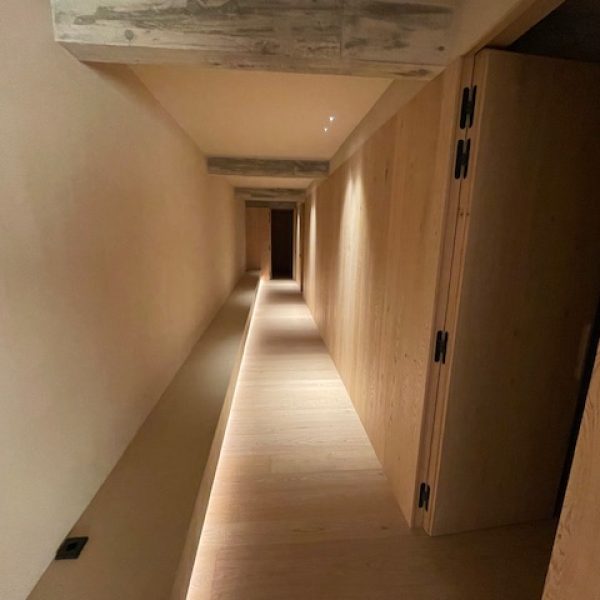 Pasillo moderno con iluminación LED en el suelo, paredes de madera clara y techo de hormigón visto, creando un ambiente cálid