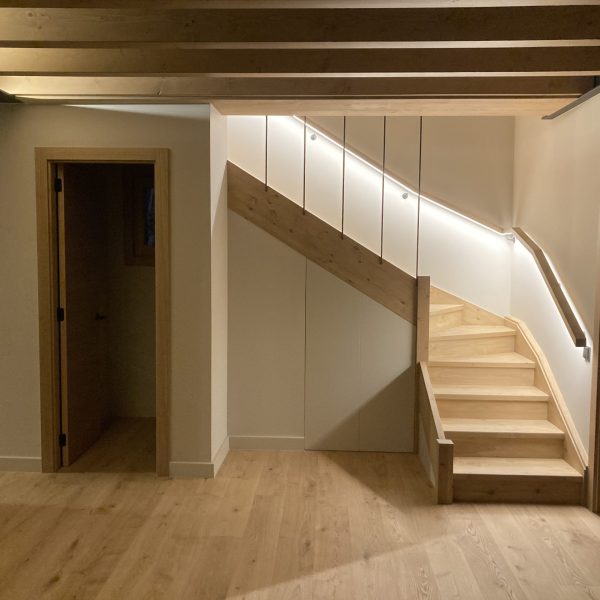 Escalera de madera clara con iluminación LED integrada en pasamanos, paredes blancas y vigas de madera en el techo.