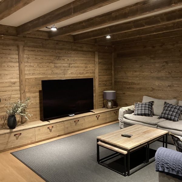 Acogedora sala de estar de estilo rústico con paredes de madera, sofá gris, TV grande y mesa de centro de madera.
