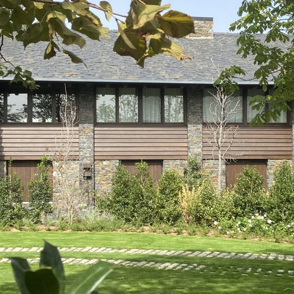 Fachada moderna de casa con revestimiento de madera, piedra y hiedra, rodeada de jardín verde y arbustos.