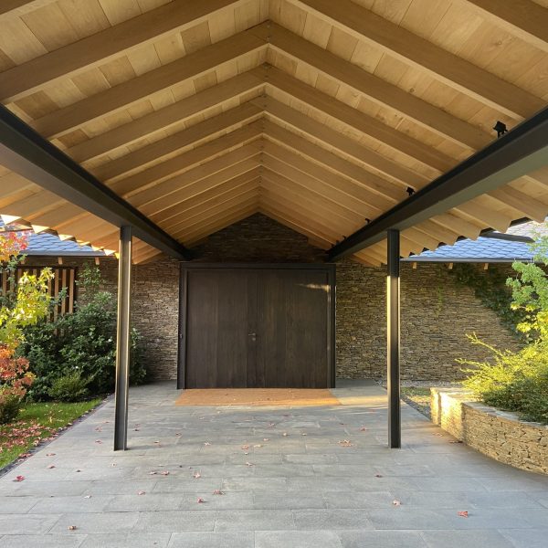 Entrada moderna a casa con techo de madera, paredes de piedra, puerta oscura y jardín otoñal con hojas rojas