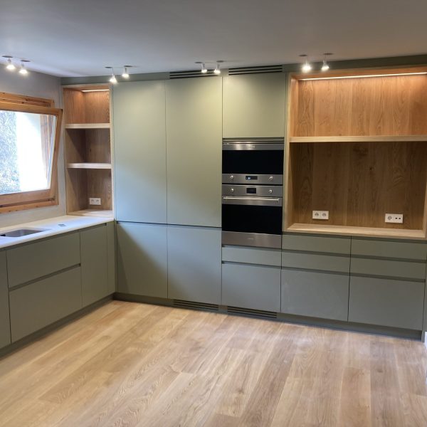 Cocina moderna con gabinetes verde salvia, encimera blanca, horno doble integrado y estantes de madera iluminados.