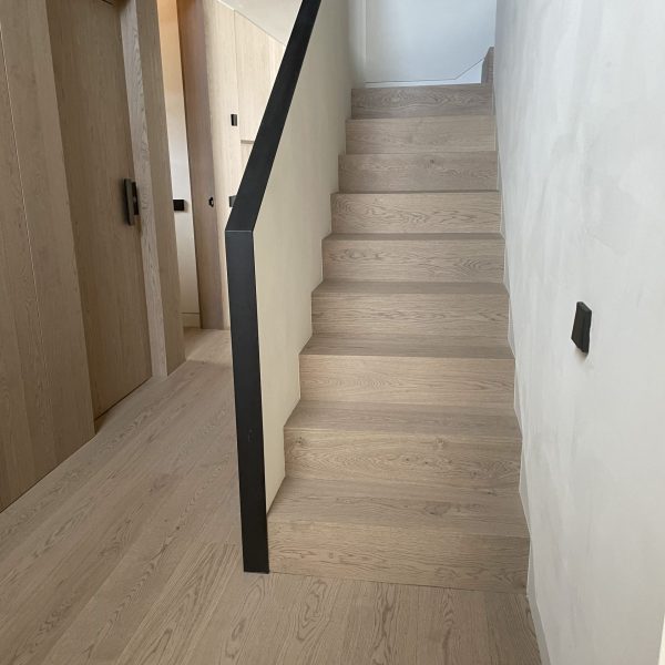 Escalera moderna de madera clara con barandilla negra minimalista y puertas de madera en un pasillo luminoso.