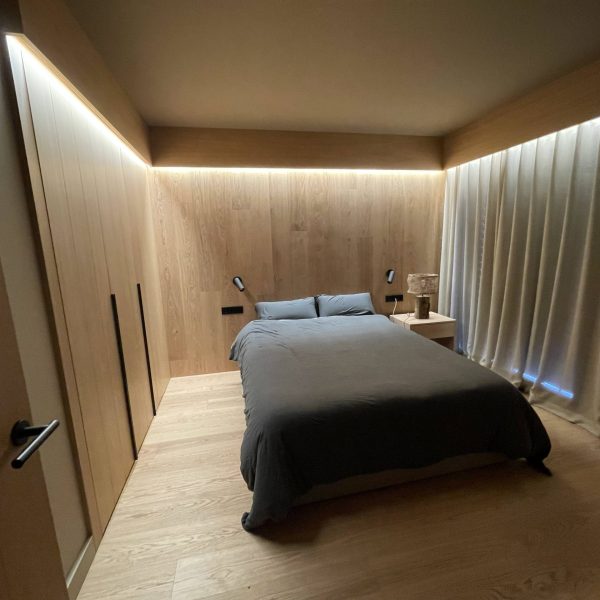 Dormitorio moderno con diseño interior de madera a medida, cama gris, iluminación LED oculta y cortinas claras