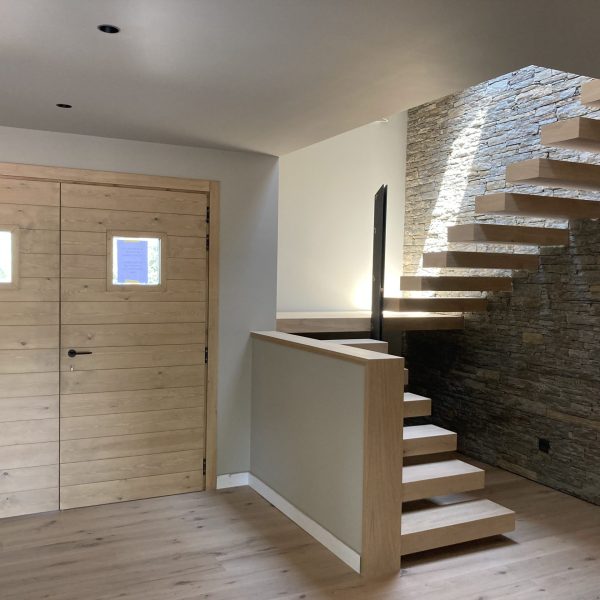 Escalera flotante de madera clara con barandilla minimalista y pared de piedra natural en un interior moderno