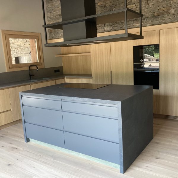 Cocina moderna con isla central gris, encimeras de piedra, gabinetes de madera clara y pared de piedra natural.