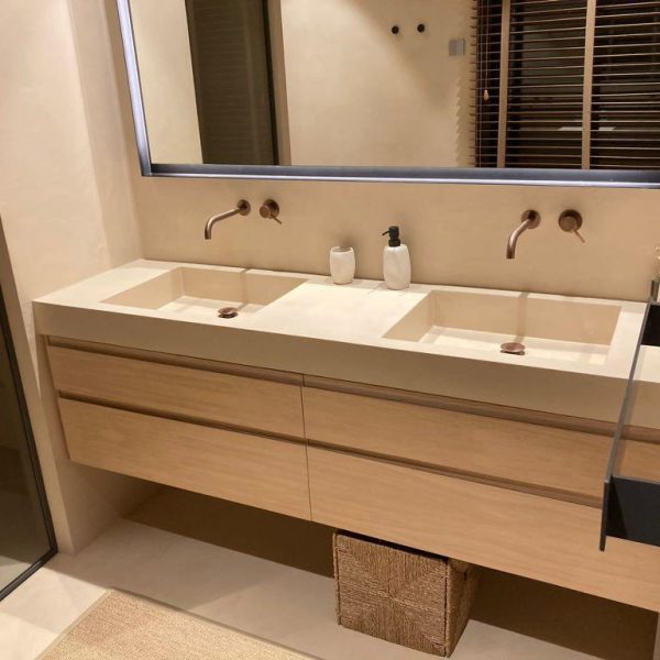 Baño moderno con doble lavabo de piedra beige, grifos de cobre en la pared y mueble de madera clara con cajones.