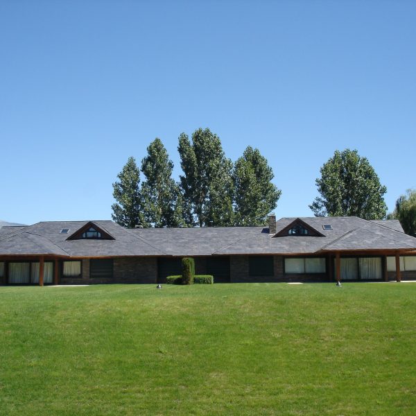 Moderna casa de campo con techo oscuro y grandes ventanales, rodeada de un extenso césped verde y árboles altos bajo un cielo
