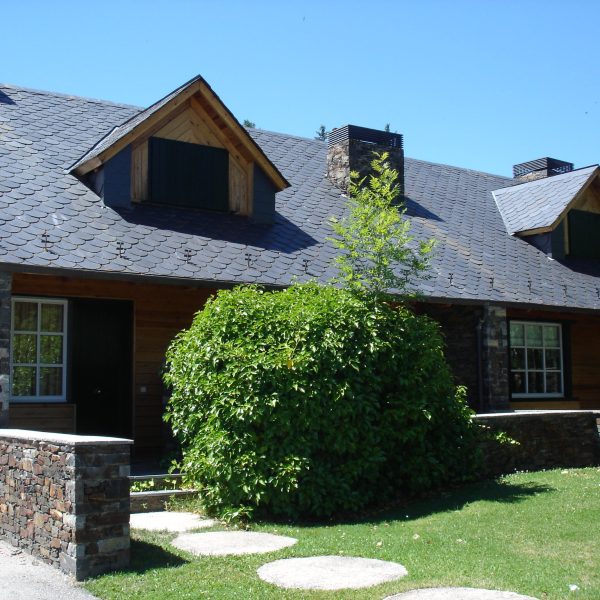 Casa de piedra y madera con tejado de pizarra, buhardillas, chimenea y jardín verde con arbustos y camino de losas.