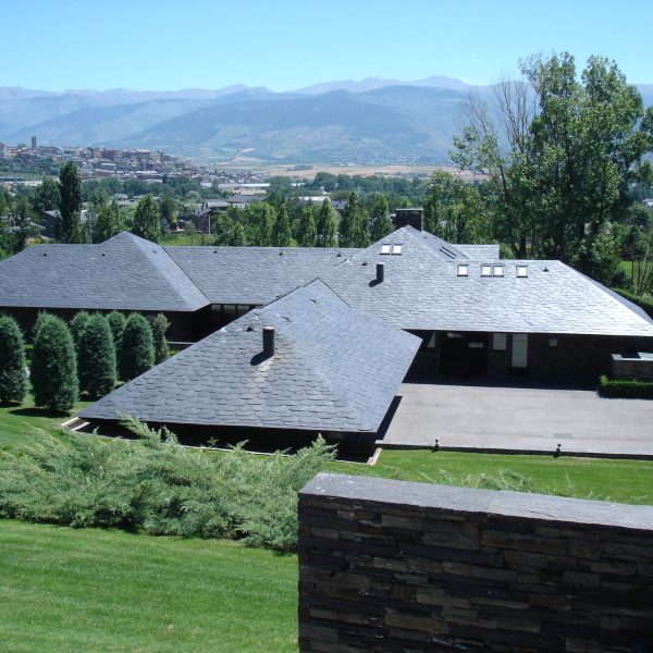Moderna casa con tejado de pizarra y jardín verde, con vistas a un pueblo y montañas en la distancia bajo un cielo azul claro