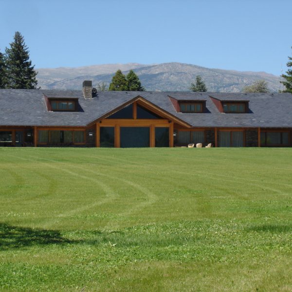 Moderna casa de montaña con tejado de pizarra, grandes ventanales de madera y amplio césped verde, con montañas al fondo.
