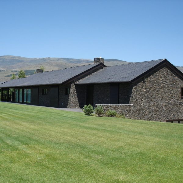 Moderna casa de piedra con tejado de pizarra y amplio césped verde, con montañas al fondo bajo un cielo azul claro.