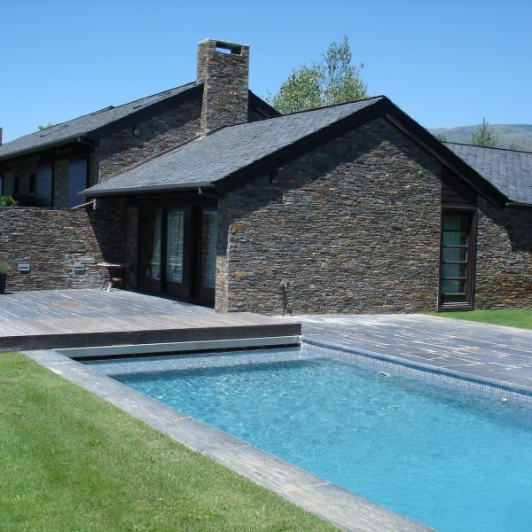 Moderna casa de piedra con piscina exterior y terraza de madera, rodeada de césped verde bajo un cielo azul claro.